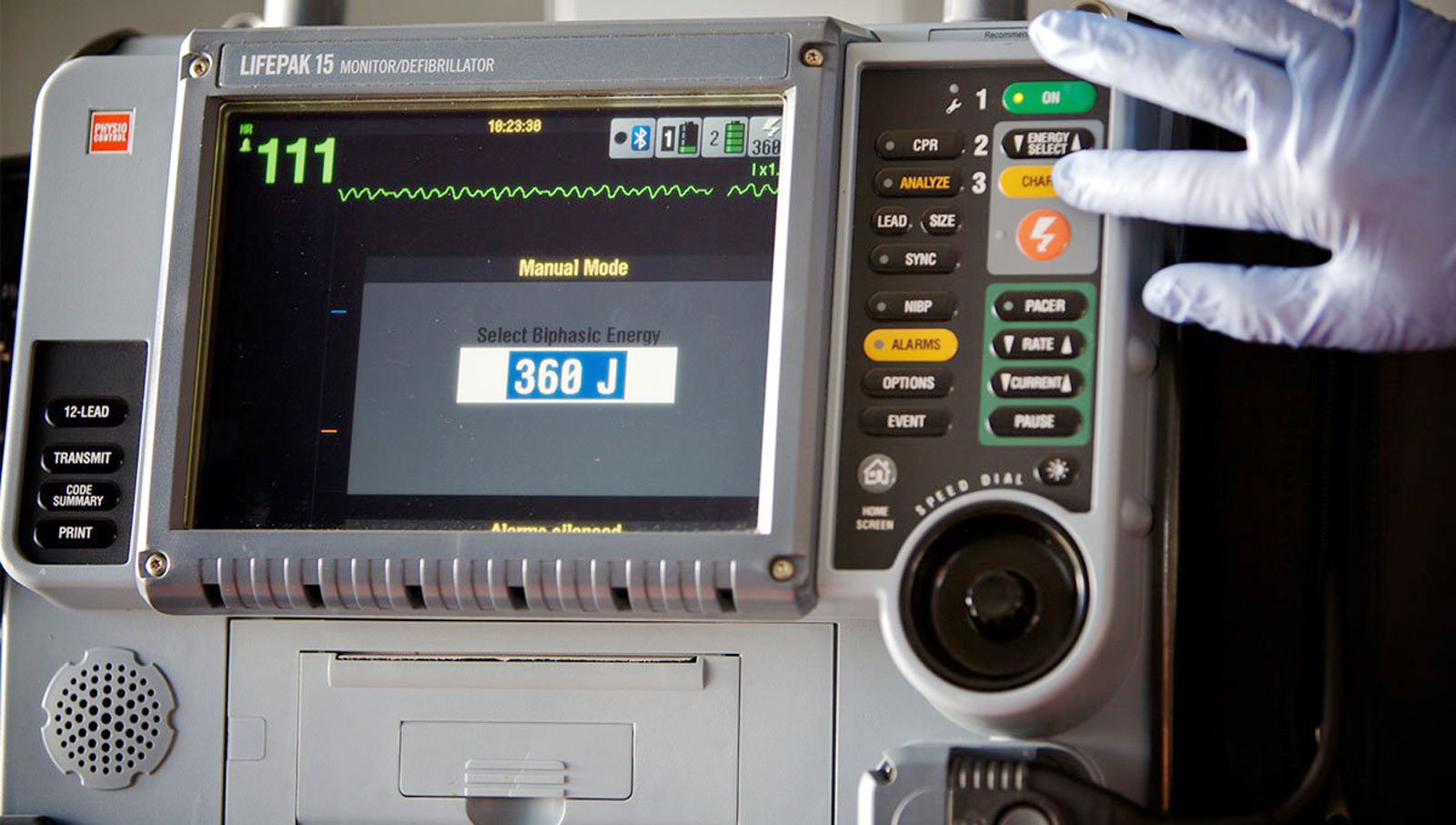 A close-up photo of the LIFEPAK® 15 defibrillator. A gloved hand presses the 'Charge' button.